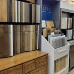 Display area showcasing various samples of wooden cabinets and drawer fronts in different finishes and styles, arranged neatly on shelves in a building supply store setting.