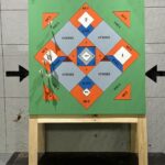 A square wooden stand holds a colorful baseball-themed target board with labeled sections for strike, ball, foul, and out—an eye-catching attraction reminiscent of axe throwing—plus arrows on a gray wall behind it.