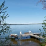 A serene resort features a calm lake with a small dock extending into the water, mooring a rowboat and a motorboat. Green foliage frames the foreground, and the horizon is clear under a bright, blue sky.