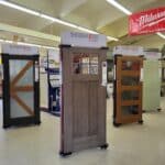 Display of four Therma-Tru doors in a store, each with different designs including glass panels. A Milwaukee tools sign is visible in the background, showcasing the quality and variety available at this building supply location.