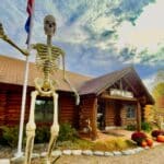 A large skeletal figure holding a flag stands proudly in front of a rustic log cabin adorned with autumn decorations, creating an intriguing display that could easily be spotlighted by the chamber of commerce.