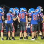 A high school football team in blue uniforms with red and white accents huddles on the field at night, discussing their next play.