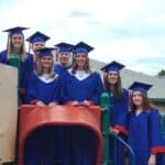 Group of six graduates in blue caps and gowns stand and pose on playground equipment, smiling at the camera.