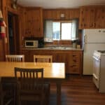 A cozy wooden kitchen in a resort, complete with a dining table and four chairs. The kitchen boasts a microwave, stove, refrigerator, sink, and various utensils. A window above the sink is adorned with charming curtains.