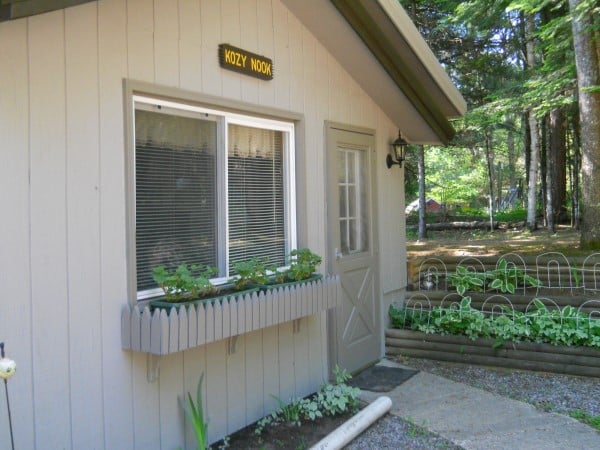 A small, light grey cabin named "Kozy Nook" with a window box of plants, a single door, and surrounded by trees and greenery.