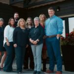 A group of eight adults poses together in front of a building, standing on a concrete walkway near a stone wall and potted plants, perhaps celebrating their teamwork in the insurance industry.