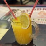 A clear pitcher filled with orange juice, garnished with a lemon slice, and two pink straws.