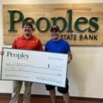 Two men stand inside Peoples State Bank, holding a large check for $500 made out to "Wounded Warrior." The check is dated September 22, 2023. One man wears a red shirt, while the other sports a blue shirt and baseball cap.