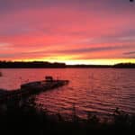 A resort sunset over a lake with a dock extending into the water, silhouetted trees on the horizon, and a vibrant sky with shades of pink, orange, and purple.