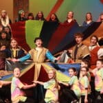 A large group of performers on stage, dressed in colorful costumes, singing with their arms outstretched and holding a multicolored fabric backdrop. Young children in tie-dye shirts are in the foreground.