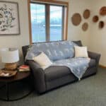 A gray couch with a blue and white patterned throw and white pillows sits in a cozy room with a round side table, a lamp, books, floral wall art, woven baskets decor on the walls, and pregnancy resource materials neatly arranged for easy access.