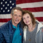 Two elderly individuals, a man and a woman, smile while embracing in front of an American flag. The man wears a blue jacket and the woman a gray sweater, embodying the warmth and unity perfect for promotional products celebrating heritage.