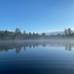 A calm lake reflects the surrounding trees and sky under a clear blue sky, with mist rising from the water's surface.