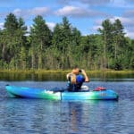 Person kayaking on a calm lake under a blue sky, surrounded by pine trees.