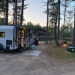 A small camper with an awning is parked in a wooded campsite near a pond. A fire pit is lit nearby, and a vehicle is parked next to the camper. Pines surround the area.