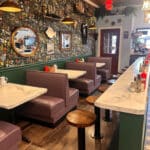 The restaurant interior features purple booth seating, marble-like tables, and a floral patterned wall. A counter with stools is perfectly complemented by various decorations and mirrors adorning the walls.