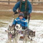 A woman and a child are enjoying sled rides on a snowy field, riding behind three running sled dogs. The woman is smiling as the child, bundled in winter clothing, marvels at the speed and strength of their huskies.