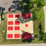 Aerial view of a sports court with red and beige markings for basketball and smaller courts, surrounded by grass, trees, an asphalt driveway leading to a parking lot with cars, and nearby buildings.