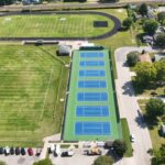 Aerial view of a sports complex featuring a running track, football field, six tennis courts, a baseball field, parking lot with asphalt driveways, and the surrounding residential area.