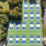 Aerial view of sixteen blue and green pickleball courts arranged in a grid on smooth asphalt, bordered by trees, a driveway, and a grassy sports field.