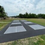 An outdoor sports area featuring eight freshly painted pickleball courts on smooth asphalt, surrounded by grass and trees under a cloudy sky, with a driveway nearby for easy access.