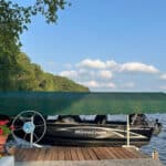 A small black MirroCraft boat sits docked under a green canopy on a wooden pier beside a lake, with potted flowers and trees in the background.
