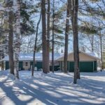 Single-story house with green siding and attached garage surrounded by tall trees and snow on a sunny winter day.