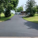 A long, newly paved asphalt driveway leads up to a two-story house with trees and a well-manicured lawn on either side. An American flag is visible near the house.