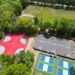 An aerial view of a recreational area featuring two outdoor basketball courts, one red and one black asphalt, surrounded by trees, and four adjacent tennis courts with blue and green surfaces. Nearby is a winding driveway leading into the park.