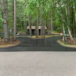 A small cabin is situated in a forest clearing with two asphalt driveways leading to it, lined with wooden fences and surrounded by tall trees.