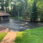 A freshly paved asphalt driveway leading to a two-car garage, surrounded by lush green grass and trees.