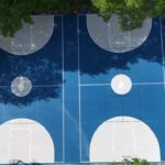 Aerial view of two blue and gray outdoor basketball courts on asphalt next to a driveway lined with parked cars, with trees casting shadows on the court.