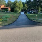 A freshly paved asphalt driveway extends from a road up to a white garage, with green trees in the background and two houses on either side. A yellow caution tape spans across the driveway entrance.