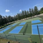 A fenced outdoor sports facility featuring six blue pickleball courts with green borders, all set against an asphalt driveway and surrounded by trees under a clear blue sky.