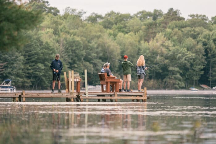 Four people are fishing from a wooden dock on a calm lake, surrounded by trees. Two stand with fishing rods, one is seated, and one watches nearby.
