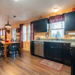 Kitchen and dining area with wood-paneled walls, dark cabinets, a wooden table with chairs, and a coffee maker on the counter. A window with checkered curtains provides natural light.