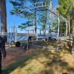 Two men work on assembling a metal frame near a lake and trees. One man in black stands on grass, while another man is partially visible within the structure.