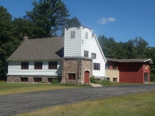 A large white house with a stone archway and chimney stands in a clearing with trees in the background, next to a red outbuilding.