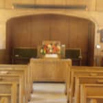 Interior of a small wooden church with rows of empty pews facing a simple altar adorned with a floral arrangement.