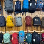 A display of assorted backpacks in various colors and styles hung on a wooden wall in a store. Tags are attached to the bags.
