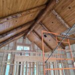 Construction site with wooden ceiling and visible beams, incomplete walls with insulation, and an orange scaffold in the foreground.