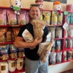 A man stands in a store holding multiple bags of popcorn gifts in front of a red shelf stocked with various popcorn products.