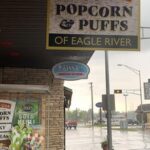 Storefront on a rainy day featuring a sign that reads "MINOCQUA POPCORN & PUFFS OF EAGLE RIVER" and an awning label for "JANS- PAINTED BIRD ICE CREAM." A red table and chair sit by the window, perfect for enjoying treats or picking up delightful popcorn gifts.