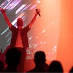 A performer with a microphone raises their arms on stage, lit by dramatic red and white lighting, with an abstract backdrop. Audience members are visible in the foreground.