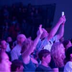 People standing in a crowded indoor venue, with someone holding up a phone to take a photo or video, while others are watching and raising their hands.