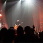 A band performs on stage with guitars and a keyboard, under bright spotlights, as an audience watches.