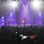 A musician plays guitar and sings on stage with band members under bright spotlights. A large audience watches the performance in an indoor venue.