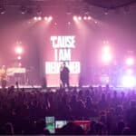 A singer performs on stage with a band, backlit by bright lights. A sign behind reads "'CAUSE I AM REDEEMED." The audience watches with raised hands.