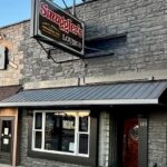 The exterior of Smuggler’s Lounge, a stone building with a large sign above the entrance and a window displaying a “Lite On Tap” sign. The door has the number 123.