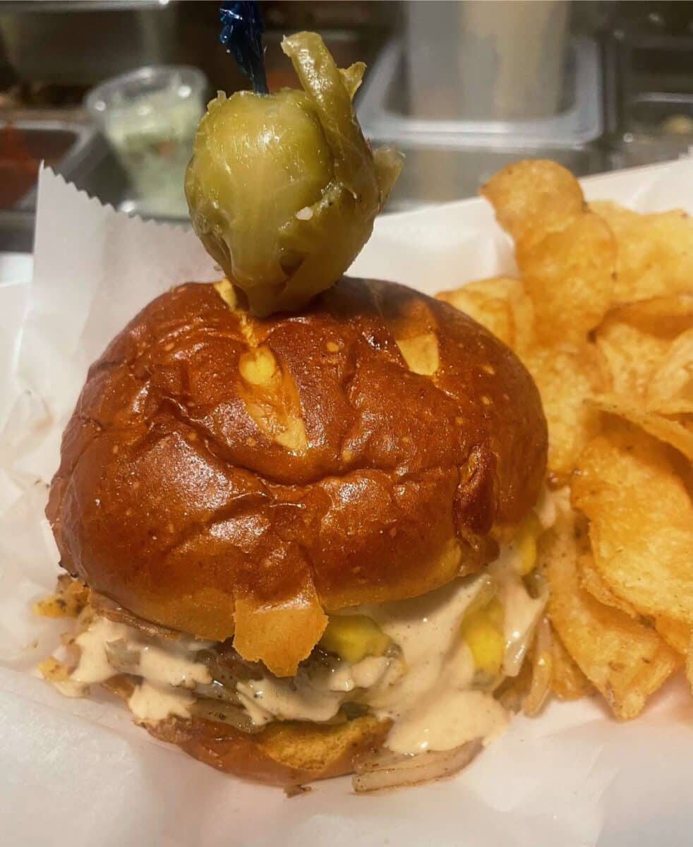A burger with melted cheese, sauce, and toppings, garnished with a pickled jalapeño on top of a pretzel bun, served alongside a portion of potato chips.